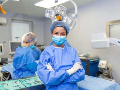 portrait-smiling-surgeon-hospital-female-healthcare-worker-is-wearing-scrubs-she-is-standing-with-arms-crossed-against-lights