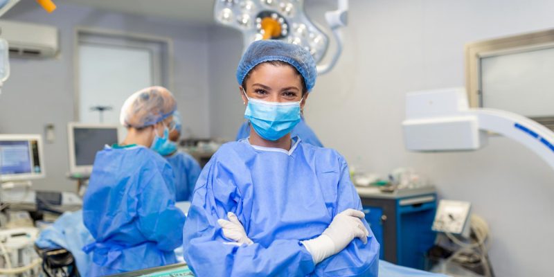 portrait-smiling-surgeon-hospital-female-healthcare-worker-is-wearing-scrubs-she-is-standing-with-arms-crossed-against-lights